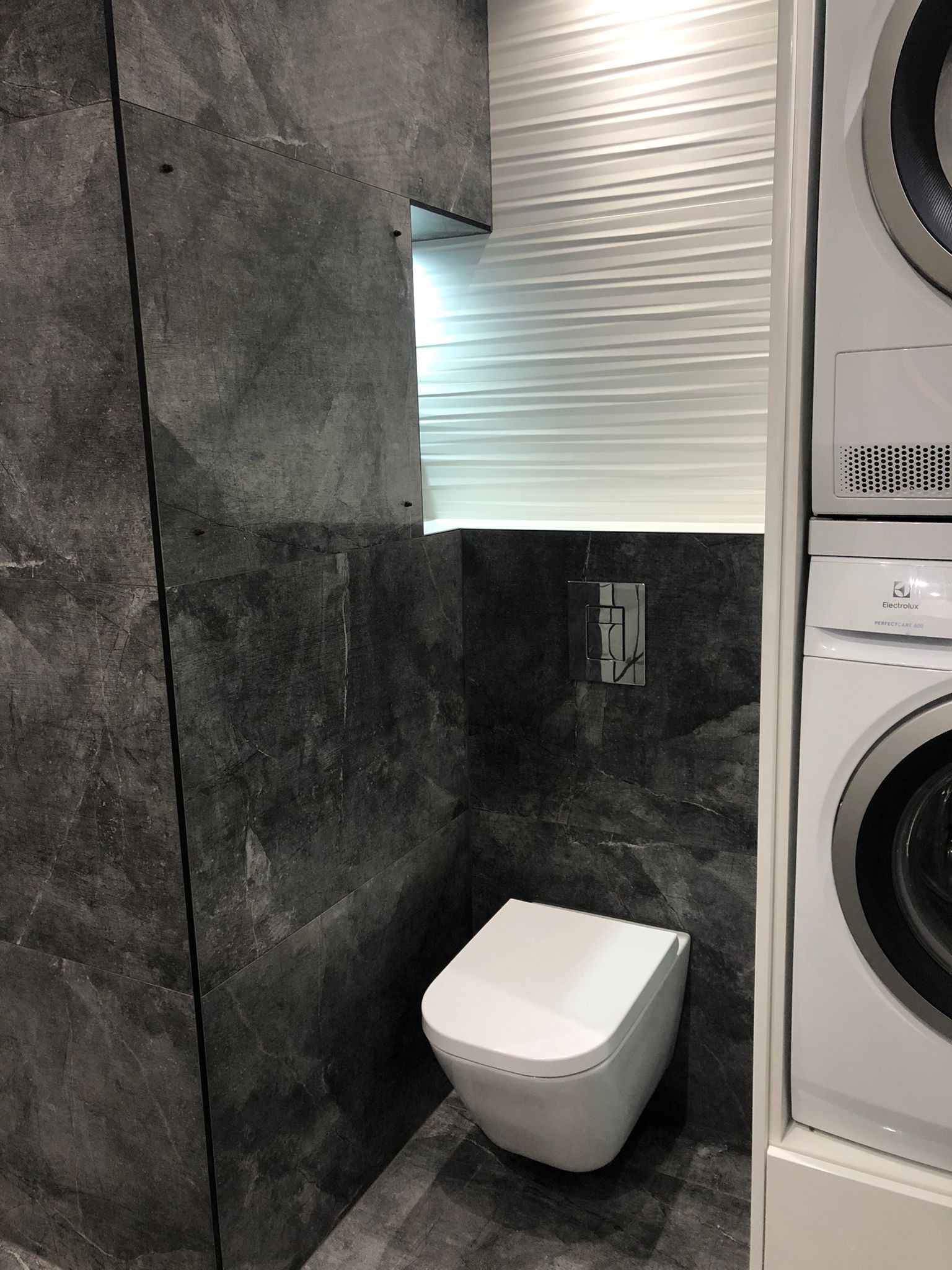 Wykończona łazienka w Legnicy z szarymi płytkami imitującymi kamień, podwieszana toaleta i pralka z suszarką w zabudowie.