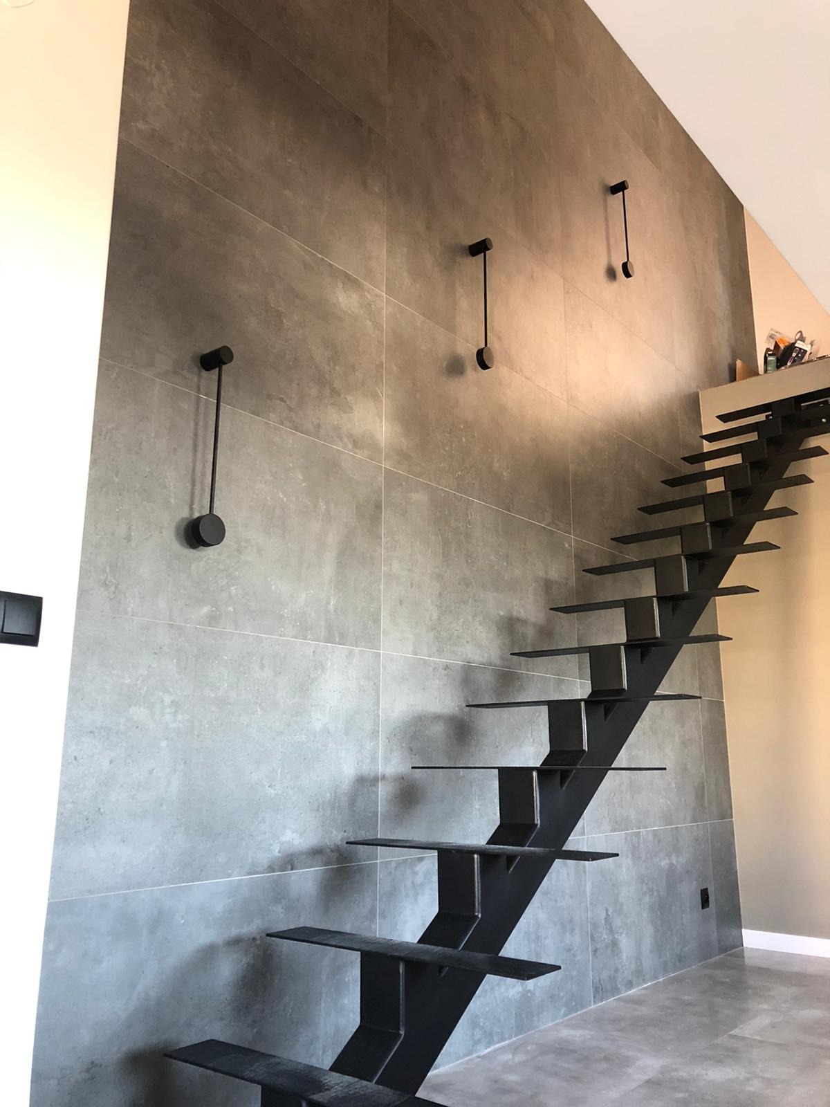 Nowoczesne schody wspornikowe w kolorze czarnym, zamontowane przy ścianie wyłożonej szarymi płytami wielkoformatowymi z minimalistycznymi, czarnymi lampami ściennymi.