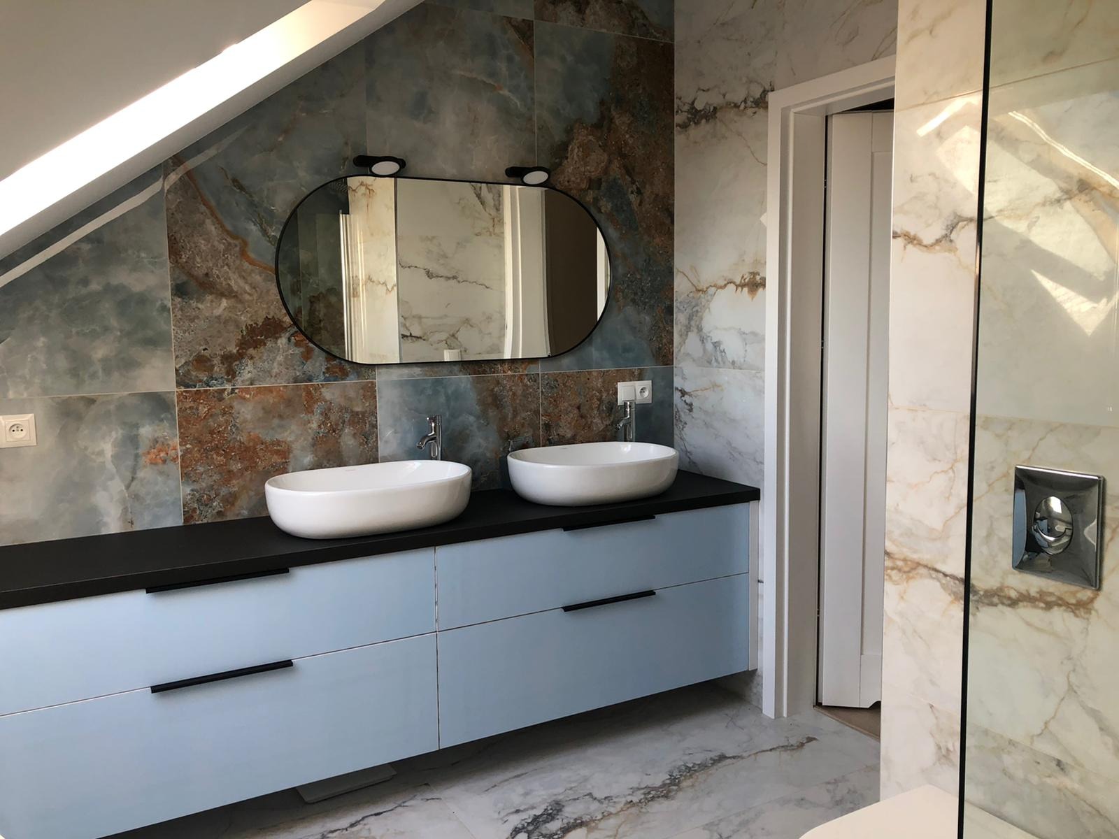 Nowoczesna łazienka na poddaszu z dwoma owalnymi umywalkami nablatowymi, szafką w kolorze gołębim i dużym lustrem w czarnej ramie na tle marmurowych płytek w odcieniach błękitu i brązu.