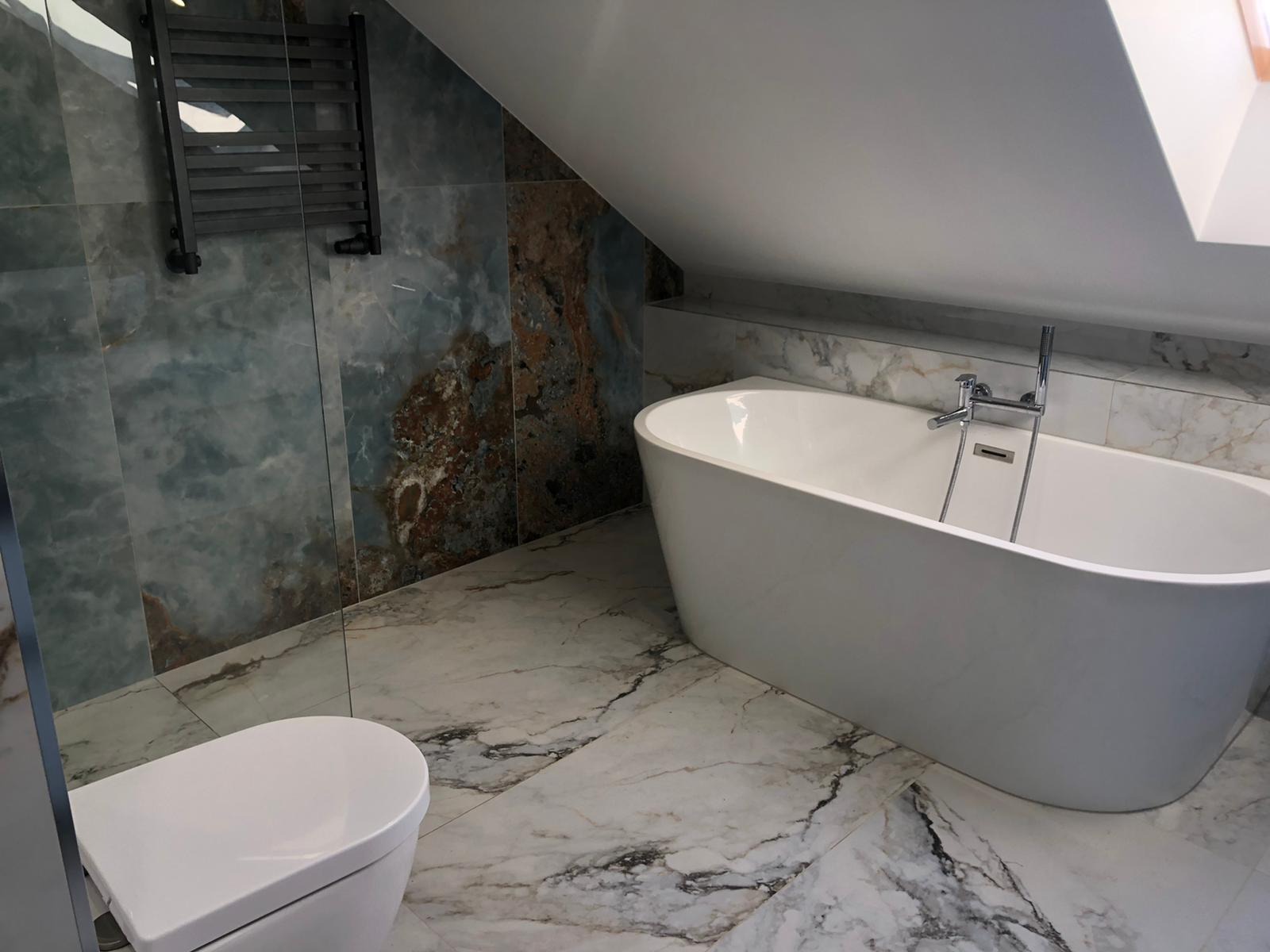 Nowoczesna łazienka na poddaszu z wolnostojącą wanną, toaletą i grzejnikiem drabinkowym na tle ściany wyłożonej płytkami imitującymi onyks.