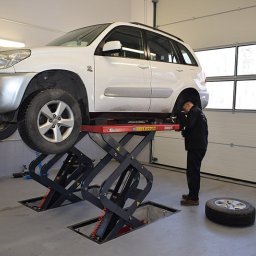 WULKAN S.C. M. Pasoń, G. Jabłoński - Biały samochód SUV na podnośniku nożycowym w warsztacie, koło leży obok, mechanik pochyla się nad podwoziem pojazdu.
