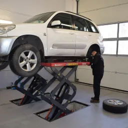 Biały samochód SUV na podnośniku nożycowym w warsztacie, koło leży obok, mechanik pochyla się nad podwoziem pojazdu.