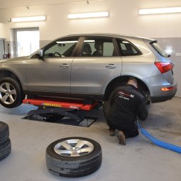 WULKAN S.C. M. Pasoń, G. Jabłoński - Srebrny SUV na podnośniku w warsztacie, pracownik w kombinezonie sprawdza zawieszenie, obok leżą zdjęte koła.