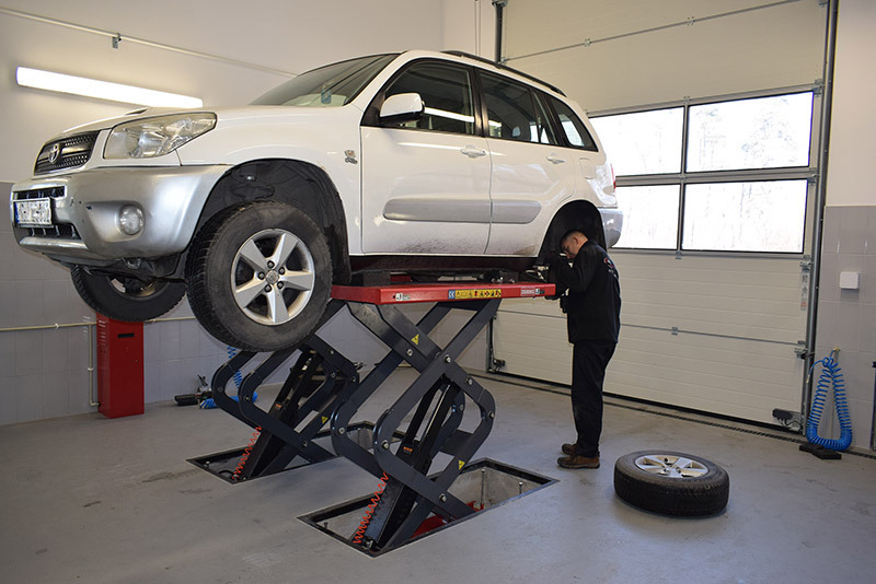 Biały samochód SUV na podnośniku nożycowym w warsztacie, koło leży obok, mechanik pochyla się nad podwoziem pojazdu.