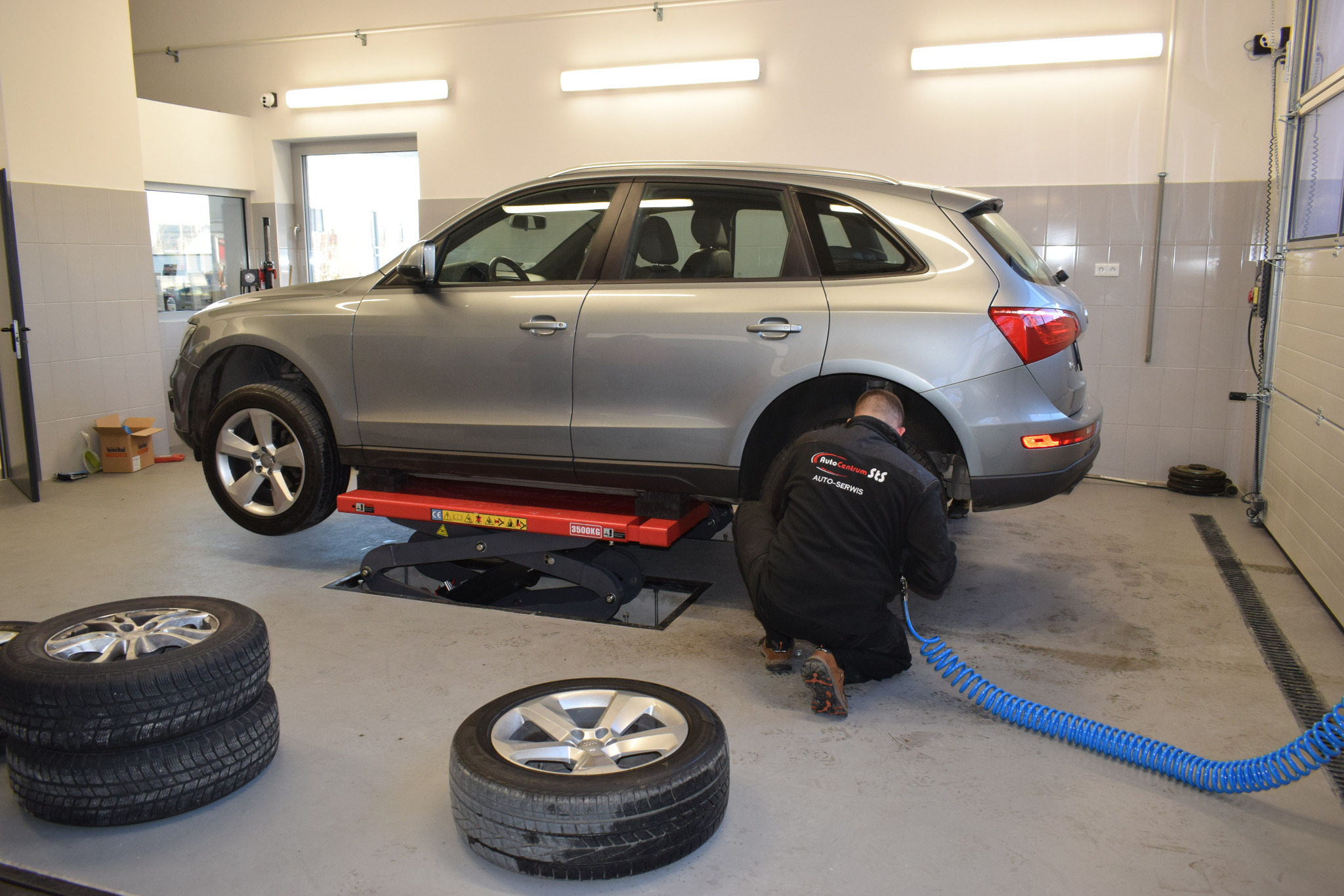 Srebrny SUV na podnośniku w warsztacie, pracownik w kombinezonie sprawdza zawieszenie, obok leżą zdjęte koła.