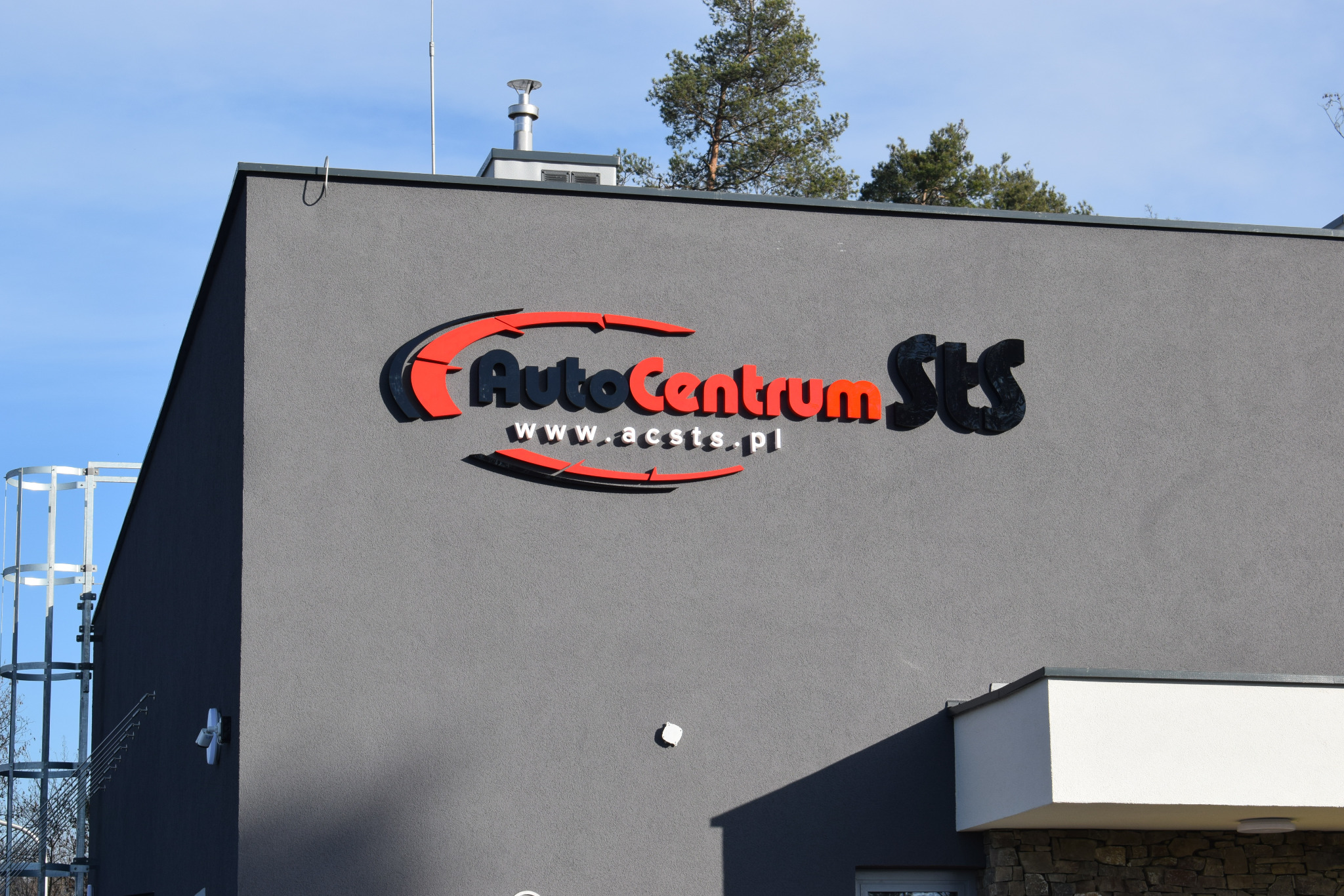 Budynek warsztatu samochodowego z logo AutoCentrum STS w kolorach czerwonym i czarnym oraz adresem strony internetowej na szarej fasadzie, widoczne drzewa w tle.