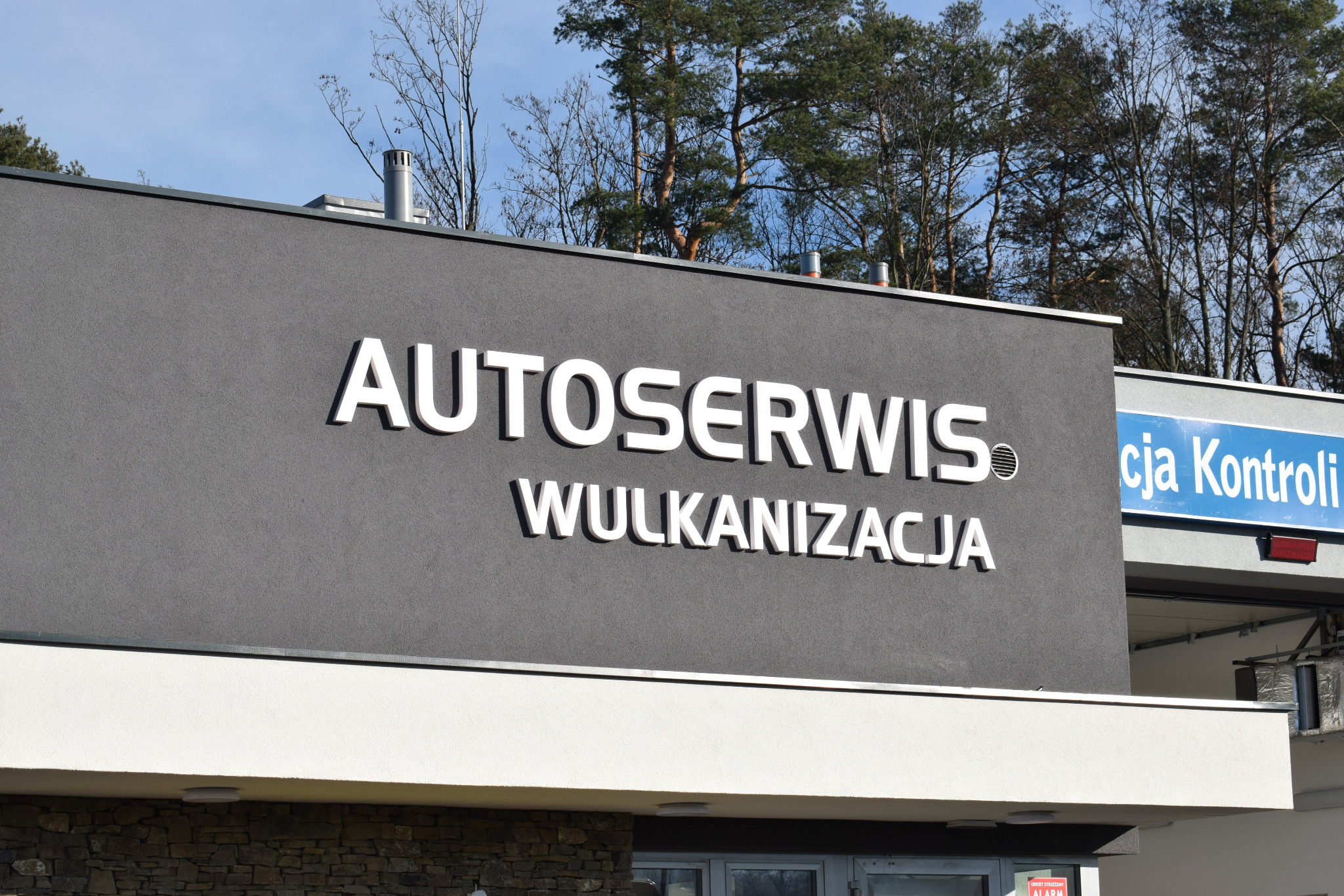 Budynek autoserwisu z napisem 'AUTOSERWIS WULKANIZACJA' na szarej ścianie, widoczne drzewa w tle i fragment tablicy 'cja Kontroli'.