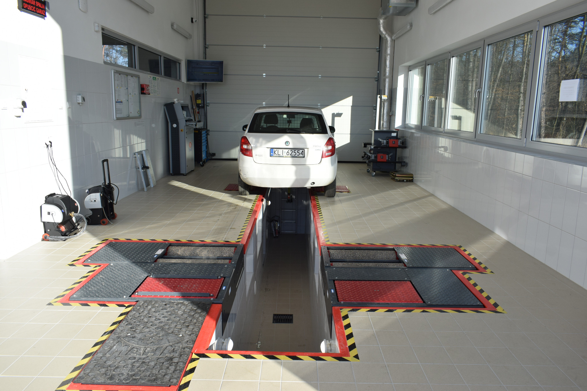 Biały samochód osobowy na stanowisku diagnostycznym w jasnym warsztacie. Widoczny kanał rewizyjny z podnośnikami i osprzęt diagnostyczny.