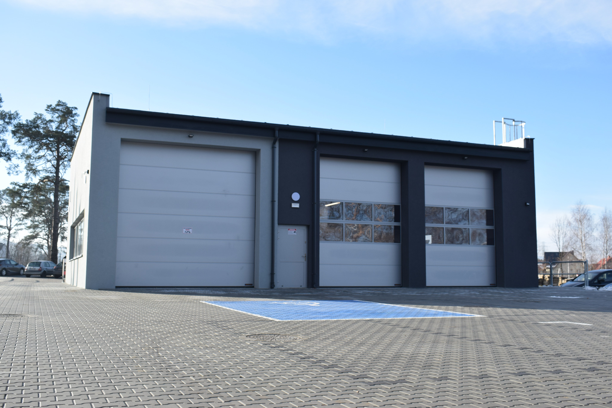Nowoczesny warsztat samochodowy z trzema bramami garażowymi i niebieskim miejscem parkingowym dla osób niepełnosprawnych na brukowanym parkingu w słoneczny dzień.