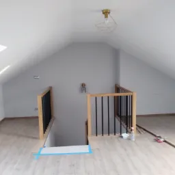 Adaptacja poddasza: drewniane panele podłogowe ułożone w jasnym pokoju na poddaszu, widoczne schody z drewnianą balustradą z czarnymi szczeblami, okna dachowe w suficie, surowa ściana pomalowana...