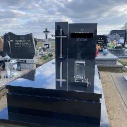 Czarny, polerowany nagrobek z krzyżem i lampionem na cmentarzu, odbijający chmury na powierzchni kamienia.