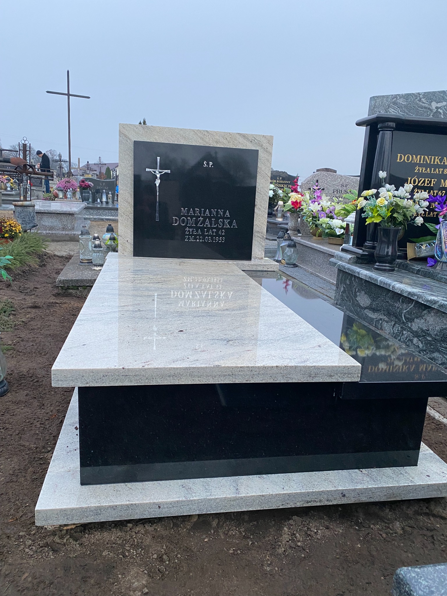 Nowoczesny nagrobek z jasnego kamienia z czarną tablicą inskrypcyjną na cmentarzu, widoczne inne groby i krzyż w tle.