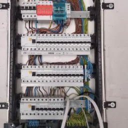 Otwarta skrzynka rozdzielcza z widocznymi bezpiecznikami, zabezpieczeniami przepięciowymi w kolorze czerwonym i niebieskim, oraz licznymi przewodami w różnych kolorach, zamontowana na szarej ścianie.