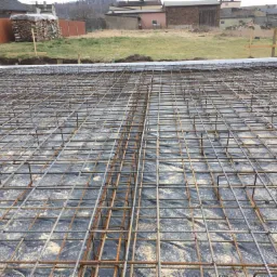 Zbrojenie fundamentu na czarnej folii, z białym styropianem na obrzeżach, przygotowane do wylania betonu, w tle zabudowa wiejska.