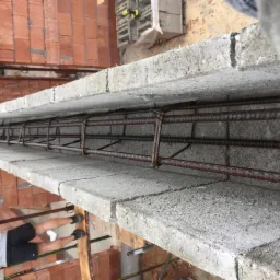 Wieniec żelbetowy w trakcie wykonywania, z widocznym zbrojeniem i betonowymi bloczkami, w tle ceglana ściana.