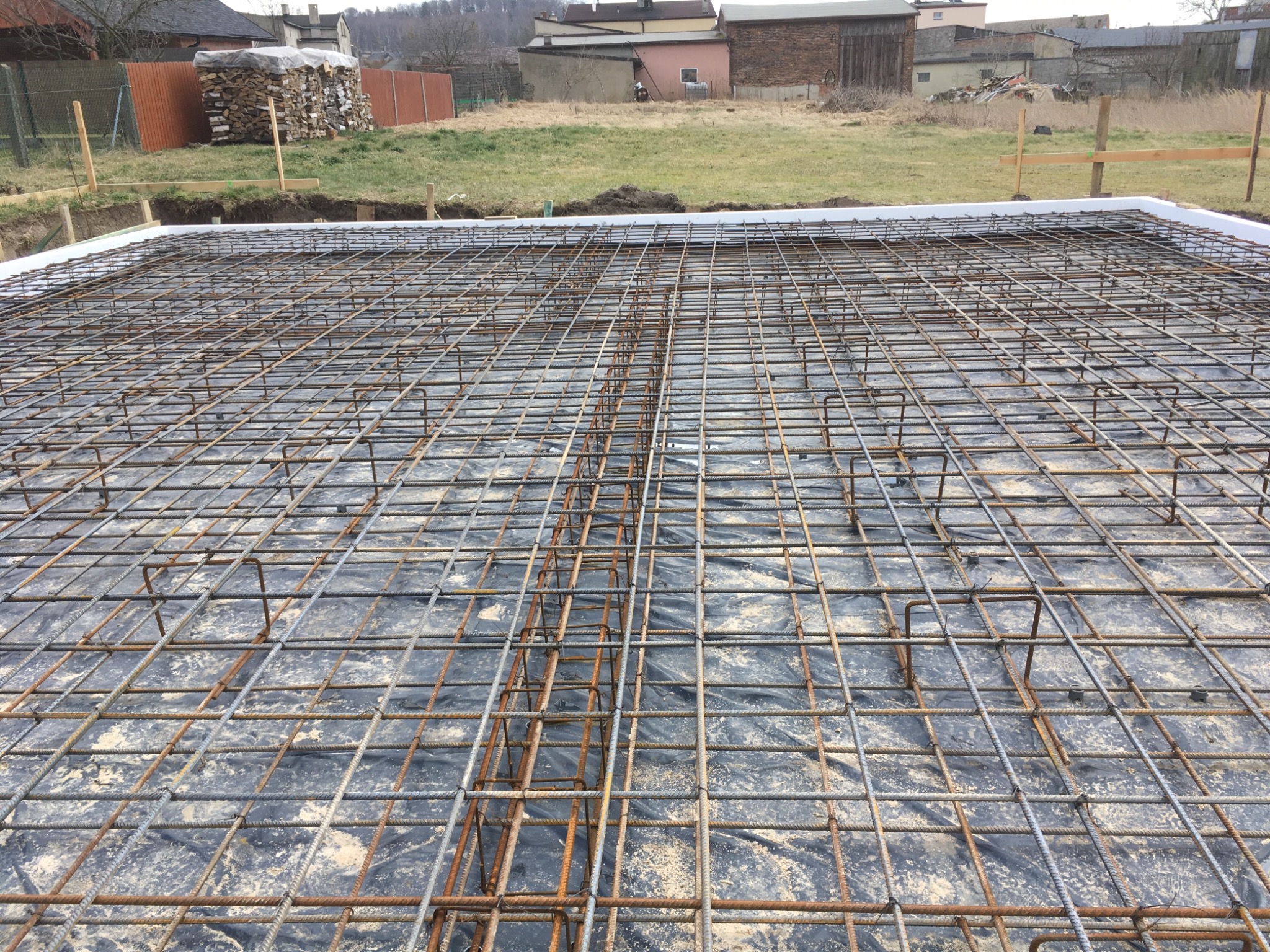 Zbrojenie fundamentu na czarnej folii, z białym styropianem na obrzeżach, przygotowane do wylania betonu, w tle zabudowa wiejska.
