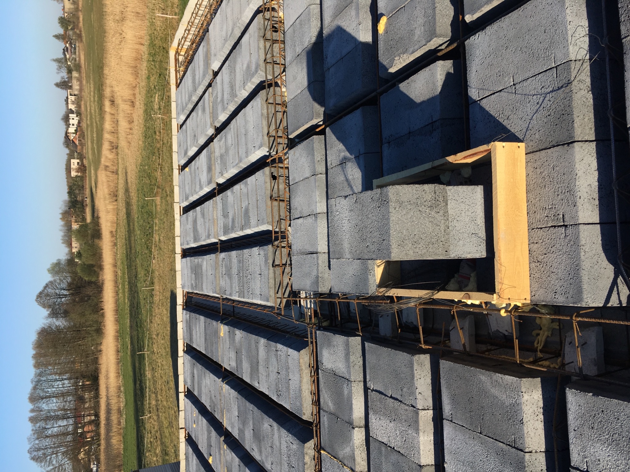 Wznoszone ściany z szarych bloczków betonowych z widocznym zbrojeniem, w tle krajobraz z polami i zabudowaniami.