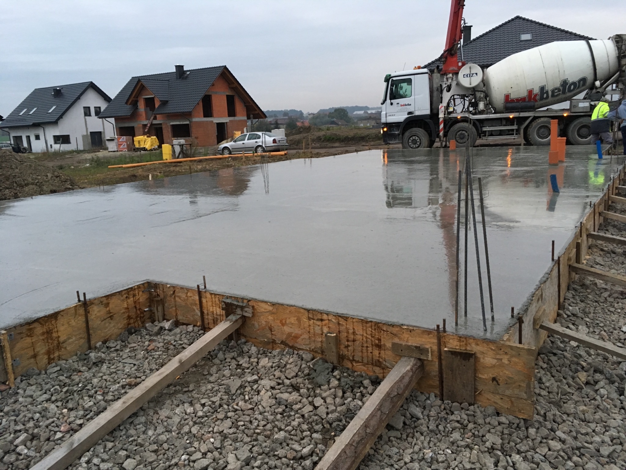 Wylany fundament pod budowę domu jednorodzinnego, widoczny betonowy plac w drewnianym szalunku, w tle betoniarka i budynki w trakcie budowy.
