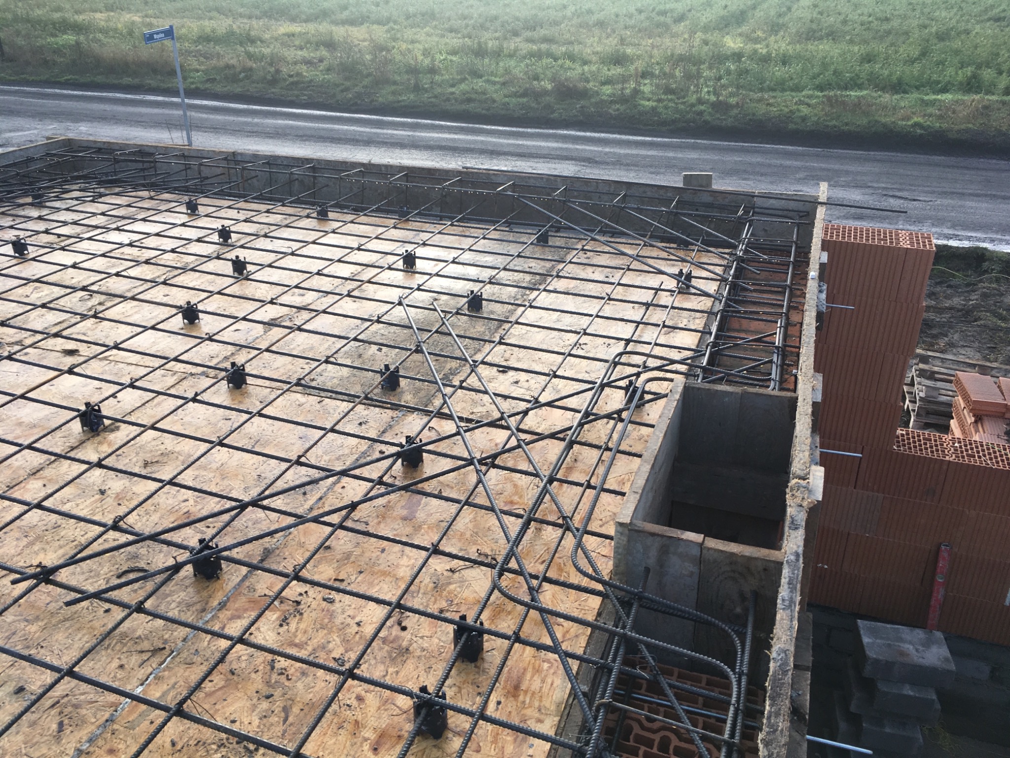 Zbrojenie płyty fundamentowej z dystansami na drewnianym szalunku, widok z góry, częściowo wylane ściany fundamentowe i paleta z pustakami ceramicznymi w tle.