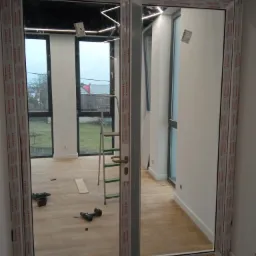 Dwuskrzydłowe drzwi balkonowe z białymi ramami, widoczne narzędzia i drabina w nowym pomieszczeniu z drewnianą podłogą, widok na zewnątrz przez okna.