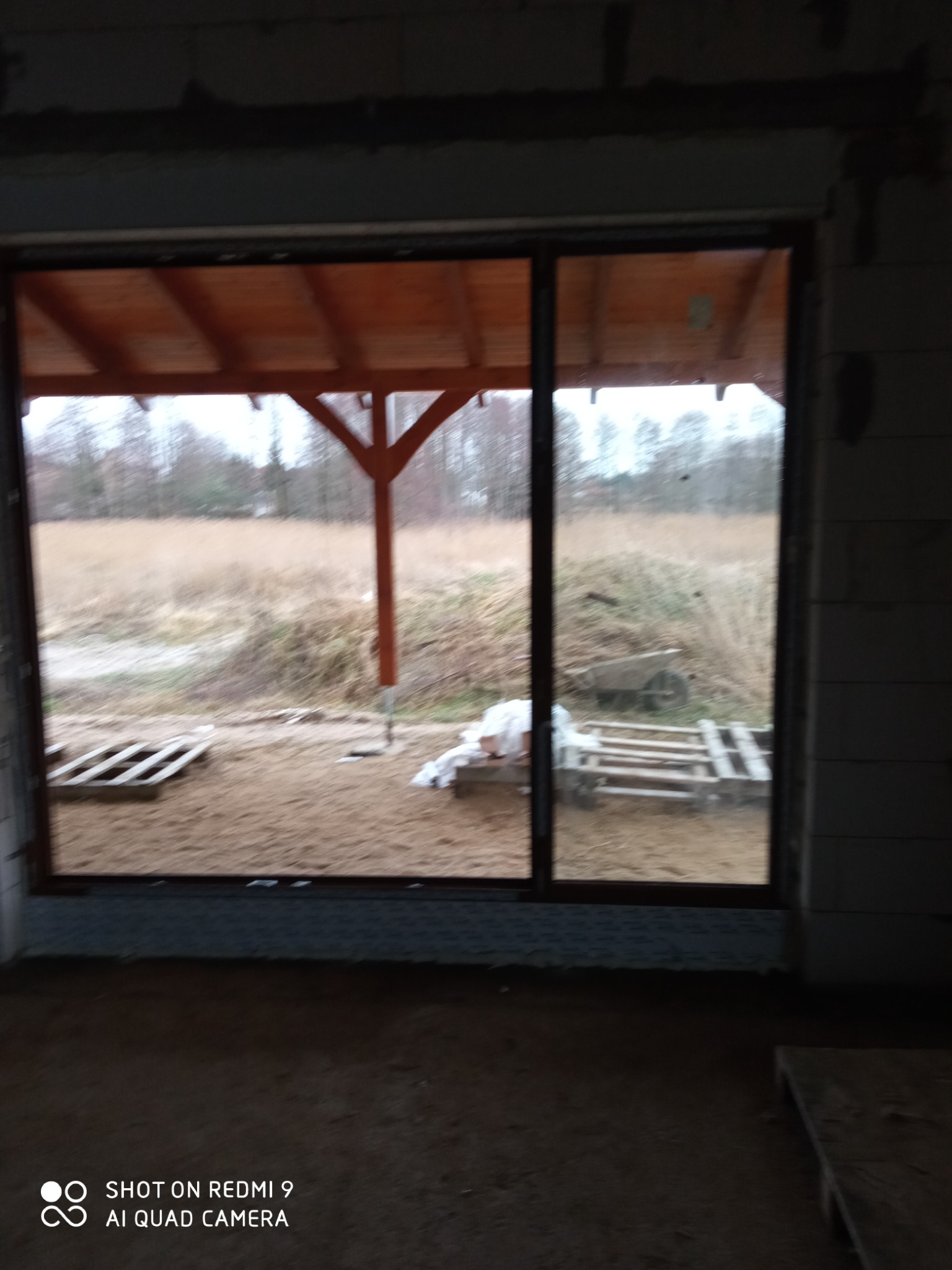 Duże przesuwne okno z widokiem na zewnątrz w surowym stanie, z widocznym drewnianym zadaszeniem na zewnątrz i paletami na ziemi, sfotografowane z wnętrza budynku w trakcie budowy.