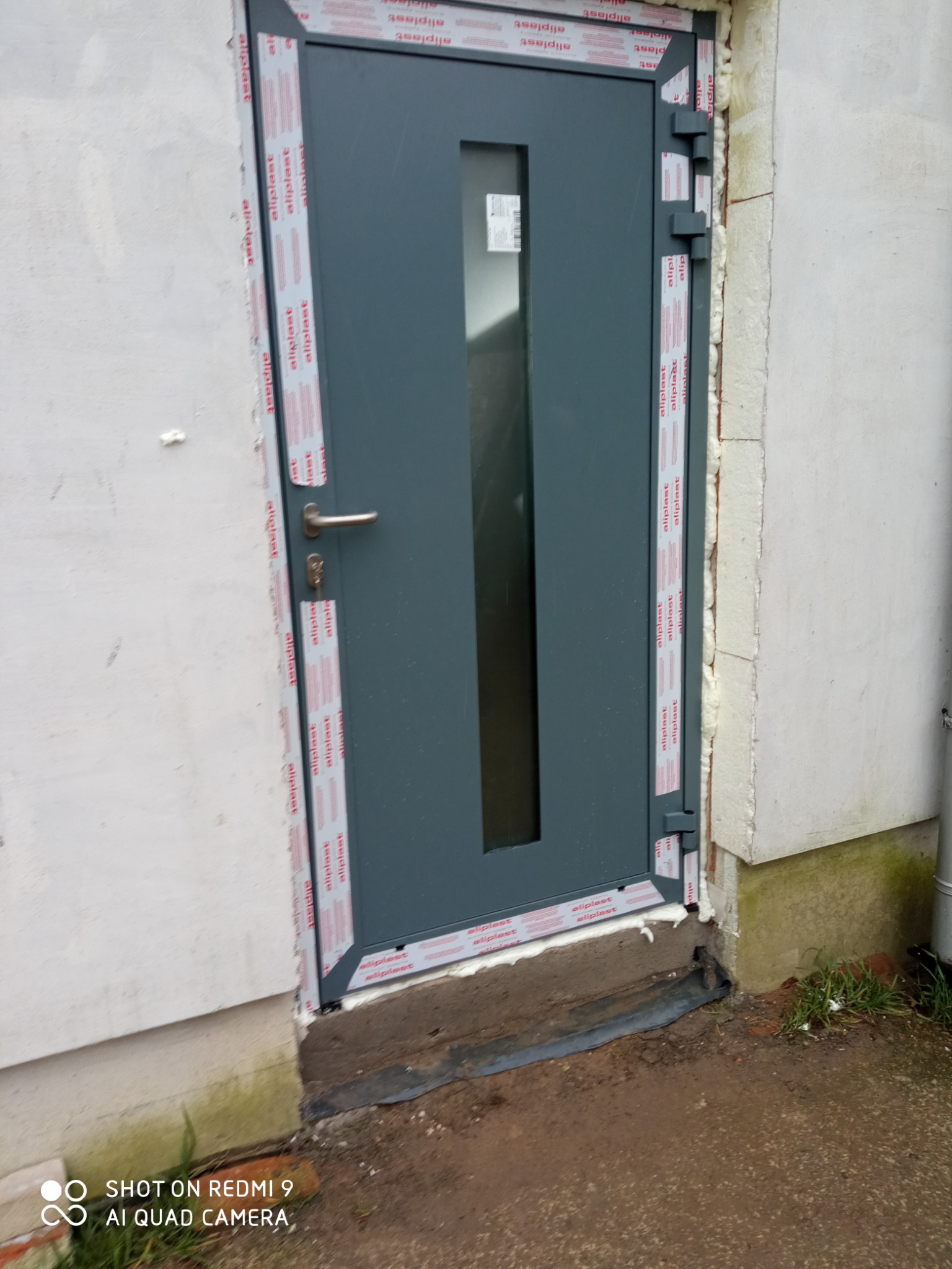 Szare drzwi wejściowe z małym okienkiem, osadzone w murze, z widoczną pianką montażową i folią ochronną na ościeżnicy.