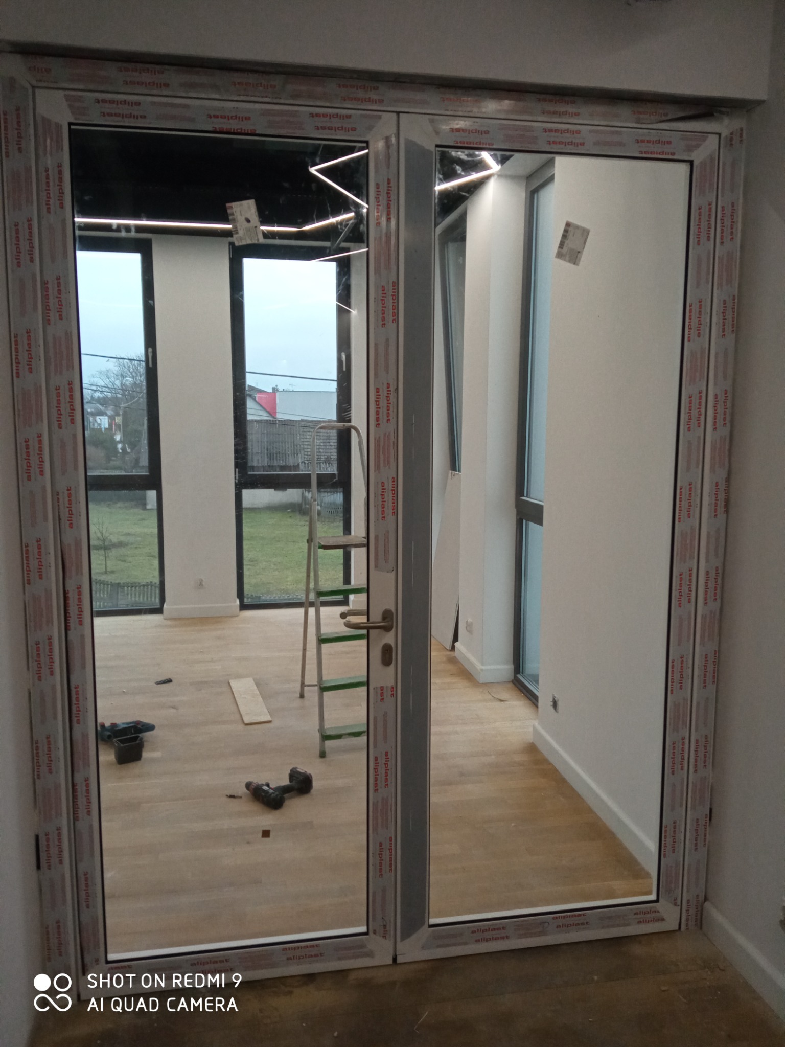 Dwuskrzydłowe drzwi balkonowe z białymi ramami, widoczne narzędzia i drabina w nowym pomieszczeniu z drewnianą podłogą, widok na zewnątrz przez okna.