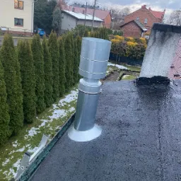 Metalowy komin wentylacyjny na dachu pokrytym papą, widok z góry, w tle rząd tuj i zabudowa jednorodzinna.