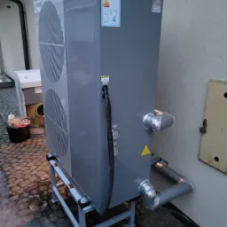 Szara pompa ciepła powietrze-woda na metalowej ramie, z widocznymi przyłączami rur owiniętymi folią aluminiową, etykietą energetyczną i czarnym kablem zasilającym.