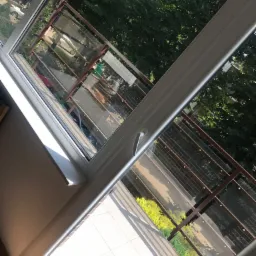 Widok z okna na balkon z przeszklonym zadaszeniem, drzewa w tle, jasne światło dzienne.