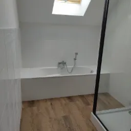 Jasna łazienka na poddaszu z drewnopodobną podłogą, białą wanną obudowaną płytkami i kabiną prysznicową z czarną ramą, doświetlona oknem dachowym.