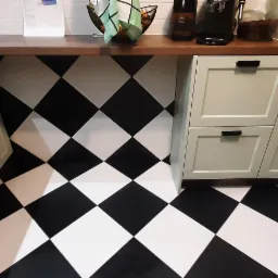 Ułożona pod kątem czarno-biała podłoga w szachownicę w kuchni z jasnymi szafkami i drewnianym blatem.