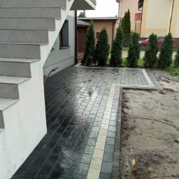 Ułożona kostka brukowa w odcieniach szarości i czerni prowadząca od schodów wejściowych do domu, zakończona rabatą z tujami; widoczne zabrudzenia po niedawnym układaniu.