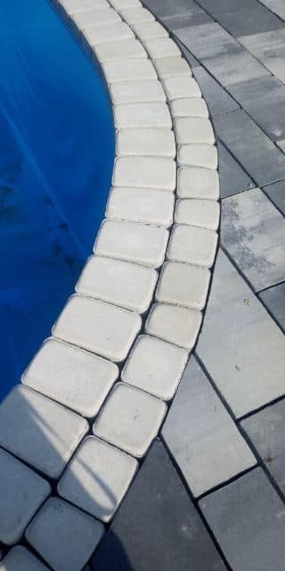 Jasne, prostokątne kostki obrzeżowe ułożone łukiem wokół niebieskiego basenu, sąsiadujące z szarym brukiem w jodełkę.