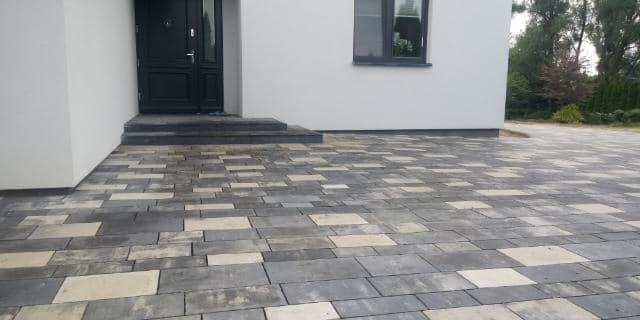 Nowoczesna nawierzchnia z kostki brukowej w odcieniach szarości i beżu przed wejściem do domu z ciemnymi drzwiami i oknem.