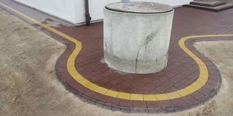 Ciemnobrązowa kostka brukowa z żółtymi akcentami ułożona wokół betonowej studzienki kanalizacyjnej, widoczne ślady po wodzie na kostce i studzience.