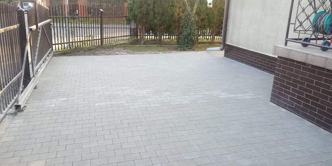 Ułożona szara kostka brukowa na podjeździe, widoczna brama wjazdowa i fragment ściany budynku z dekoracyjnym ceglanym wykończeniem.