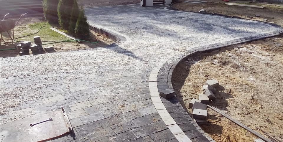 Fragment układanej nawierzchni z kostki brukowej w odcieniach szarości, widoczne przygotowane podłoże, obrzeża z kostki oddzielające różne typy nawierzchni, w tle taczka i ułożone kostki.