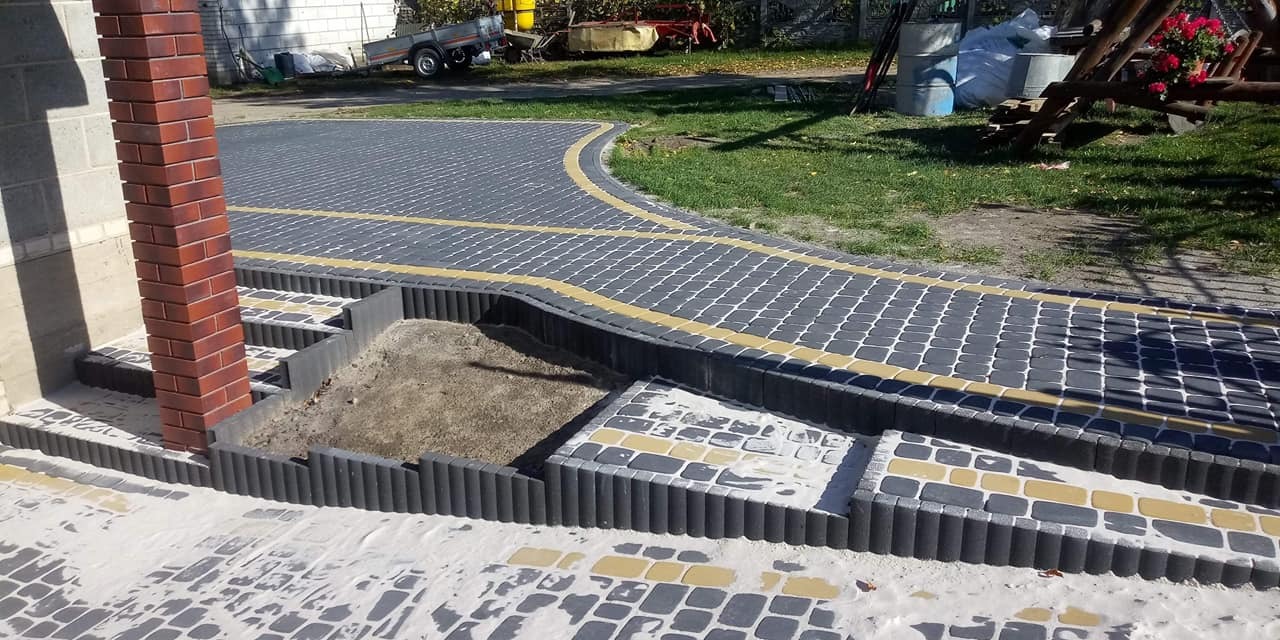 Ułożona kostka brukowa w odcieniach szarości i żółci, z obrzeżami w kształcie walców, tworząca schody i chodnik. W tle widoczny trawnik, drzewa i przyczepka.