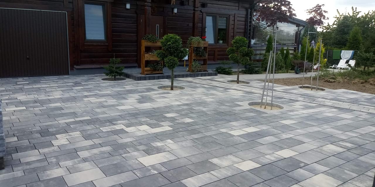 Ułożona kostka brukowa w odcieniach szarości przed drewnianym domem z garażem; w nawierzchni przygotowane okrągłe miejsca na nasadzenia drzewek.