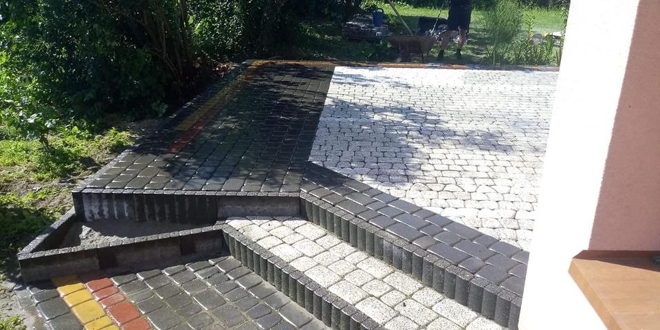 Fragment tarasu z kostki brukowej w dwóch kolorach, schody z obrzeżami, widoczne prace wykończeniowe, w tle trawnik i osoba z taczką.