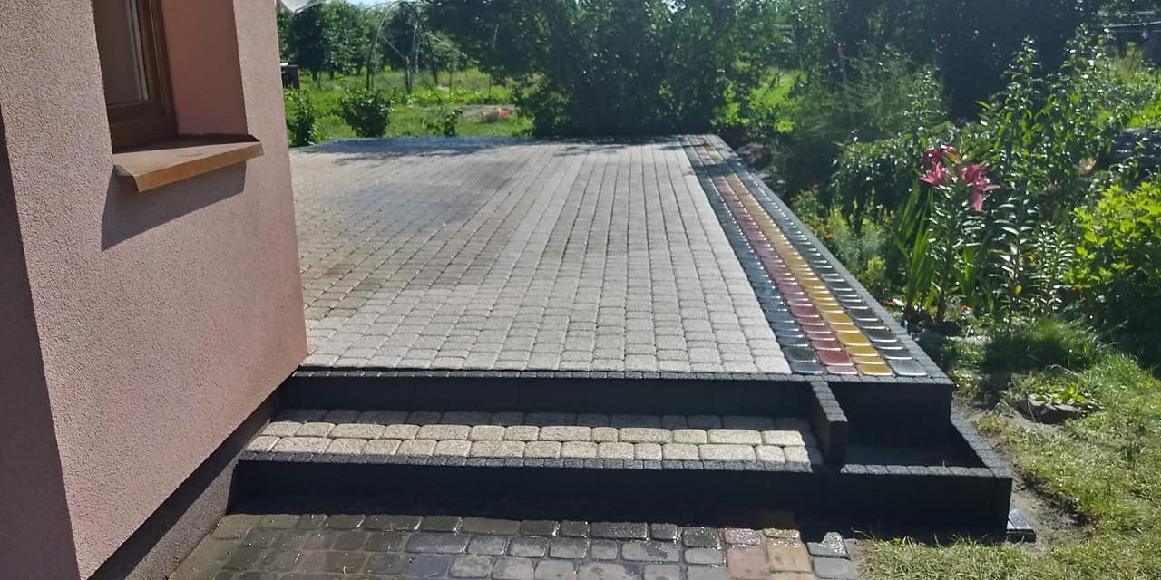 Nowo ułożona kostka brukowa w odcieniach szarości i beżu prowadząca do domu, z dekoracyjnym obrzeżem z kolorowych kostek i dwustopniowymi schodami, otoczona zielenią ogrodu.