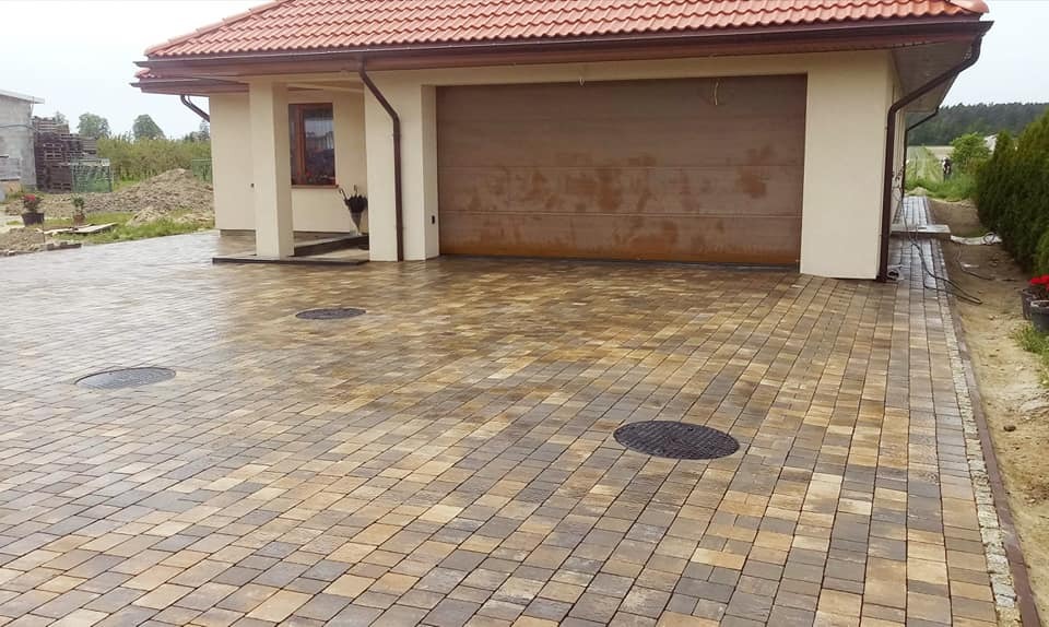 Ułożona kostka brukowa w odcieniach brązu i beżu przed domem z garażem, widoczne trzy studzienki kanalizacyjne oraz obrzeża z kostki granitowej.