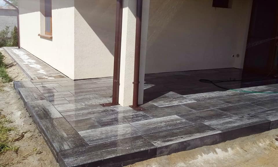 Nowo ułożony taras z szarych, prostokątnych płyt, obramowany ciemniejszymi obrzeżami, przylegający do elewacji budynku z brązowymi rynnami i oknem.