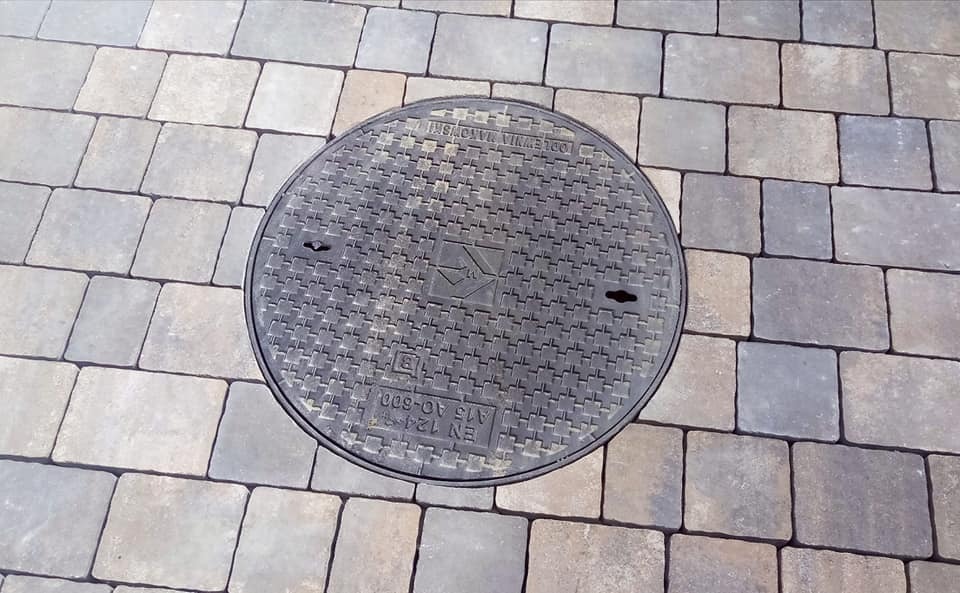 Szara kostka brukowa ułożona w regularny wzór z okrągłą, żeliwną studzienką kanalizacyjną pośrodku. Pokrywa studzienki ma wzór kratki i oznaczenia producenta oraz normy.
