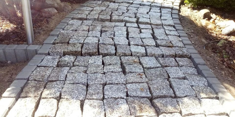 Ścieżka z nieregularnych, szarych kostek granitowych, obramowana regularnymi, szarymi obrzeżami betonowymi, prowadząca przez ogród.