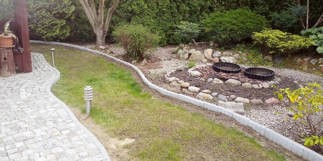 Kaskadowy ogród z obrzeżami trawnikowymi z szarej kostki, ścieżka z jasnej kostki granitowej i oświetlenie ogrodowe w tle bujna zieleń.