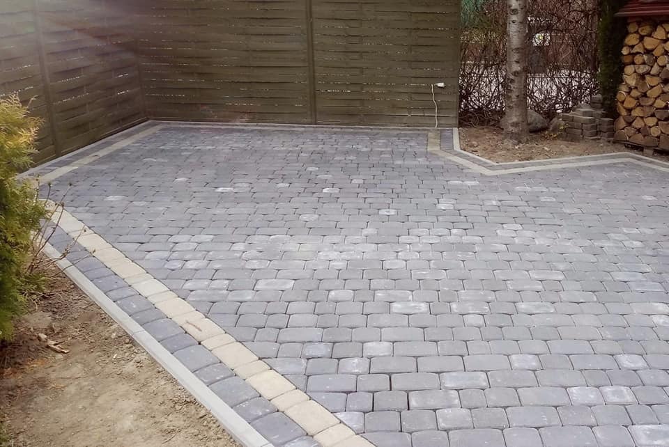 Ułożona szara kostka brukowa z jasną obwódką na posesji, widoczny fragment drewnianego ogrodzenia i drzewo w tle.
