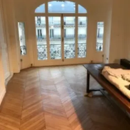 Jasne wnętrze z parkietem ułożonym we wzór jodełki francuskiej, duże okno z widokiem na miasto, częściowo widoczny stary drewniany stół.