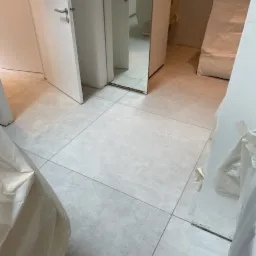 Jasnoszare, kwadratowe płytki podłogowe z widocznymi fugami, częściowo zasłonięte folią ochronną, z białymi drzwiami i lustrem w tle.